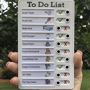 Può includere: Una lavagna bianca "To Do List" con un elenco di compiti, tra cui lavarsi i denti e rifare il letto. Ogni compito ha un segno di spunta o una X per contrassegnare il completamento. La lavagna è tenuta contro uno sfondo verde sfocato.