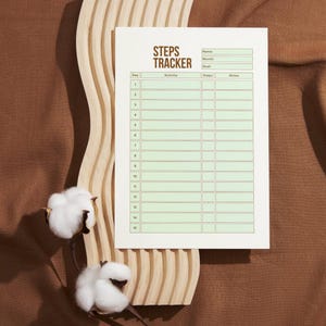 May include: A white steps tracker with green grid lines and the text "STEPS TRACKER". The tracker is on a light brown wooden decorative piece, with two cotton flowers on the left side. The background is a brown fabric.