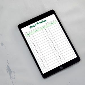 May include: A black tablet displays a "Steps Tracker" form with green headings and a white background. The form is designed to track daily steps, with columns for days, dates, and steps. The tablet is on a light gray surface.