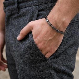 Puede incluir: Una pulsera gris oscuro con eslabones cuadrados se lleva en la muñeca. La persona lleva pantalones grises oscuros con una mano en el bolsillo. El fondo es una pared de piedra gris claro.