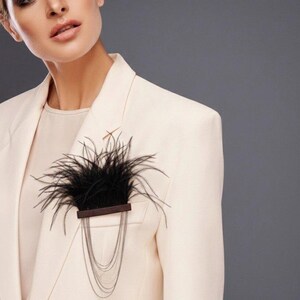 May include: A brooch featuring a horizontal dark brown bar adorned with black feathers extending upwards and multiple thin silver-toned metal chains hanging downwards in a layered, curved arrangement, pinned to a light cream-colored blazer.