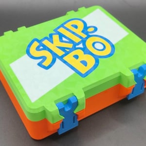 Puede incluir: Una colorida caja del juego Skip-Bo. La caja es principalmente verde con laterales naranjas y cierres azules. La parte superior presenta el logotipo del juego en amarillo y azul. La caja está cerrada y lista para jugar.
