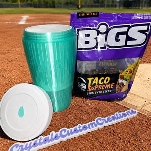 May include: A teal tumbler with a white lid and a bag of Taco Supreme sunflower seeds. The tumbler is cylindrical with a textured surface. The bag of seeds is purple with the brand name "BIGS". A white sign with the text "Crystals Custom Creations" is also present.