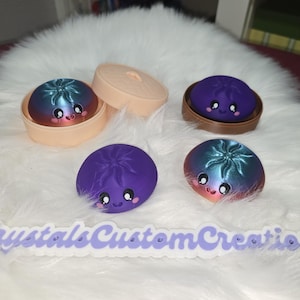 Puede incluir: Una colección de figuras en forma de dumpling sonrientes y coloridas. Algunas están en pequeños recipientes, mientras que otras se muestran individualmente. Los dumplings presentan un degradado de colores, incluyendo azul, morado y naranja, con expresiones faciales lindas. El texto "CrystalsCustomCreations" está en la parte inferior.