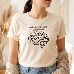 Könnte beinhalten: Cremefarbenes T-Shirt mit dem Text "not broken. not wrong. just different." und einer schwarzen Gehirngrafik. Das T-Shirt hat hochgekrempelte Ärmel. Das Model trägt eine blaue Jeans. Das T-Shirt ist aus weichem Material.