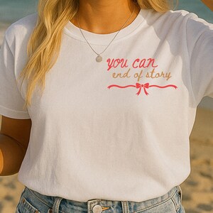 May include: White t-shirt with the text "you can end of story" in pink script and a bow design. The shirt is paired with blue denim jeans. The image is taken outdoors on a sunny day.