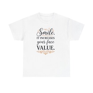 以下が含まれることがあります： 「Smile, it increases your face VALUE」というメッセージが黒と金の筆記体で書かれた白いTシャツ。クルーネックと半袖です。テキストはシャツの前面の中央に配置されています。