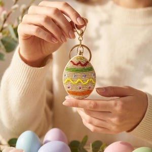 May include: A beaded Easter egg keyring with a gold-coloured ring and clasp. The egg features colourful beads in green, red, yellow, and orange. The keyring is held by a person, with several pastel-coloured Easter eggs in the background.