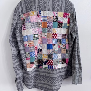 Può includere: Una camicia a maniche lunghe con bottoni in tessuto grigio con un motivo geometrico ripetitivo. Il pannello posteriore presenta un grande design patchwork rettangolare composto da numerosi piccoli quadrati di tessuto multicolore con varie stampe floreali, a pois e a quadri.