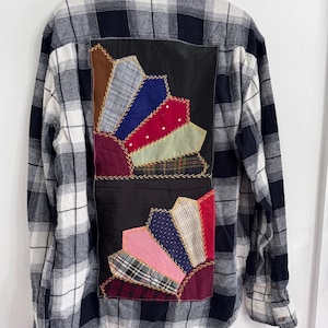 May include: A long-sleeve button-down shirt in a black, white, and gray plaid pattern. The back features two large, rectangular fabric panels appliquéd with a fan-shaped patchwork design made of various colorful fabric scraps and yellow stitching.