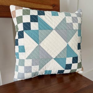 May include: Square decorative pillow with a geometric patchwork design in shades of blue, gray, and white. The pillow is quilted with a diamond pattern and rests on a wooden chair.
