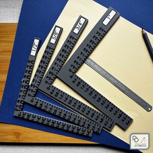 May include: A collection of gray metal speed squares and a ruler, with measurements in inches. The squares are labeled with sizes from 1/2" to 1". The tools are arranged on a light brown surface, with a blue and wooden background.