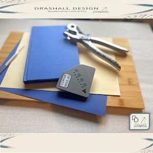 Può includere: Un insieme di strumenti e materiali per la rilegatura. Include una perforatrice argentata, una dima angolare grigia con la scritta "CORNER HOLE JIG", un libro blu e fogli di carta su una tavola di legno. Marchio "DRASHALL DESIGN".