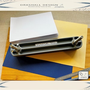 May include: A grey signature press, labelled "SIGNATURE PRESS", rests on a wooden surface with blue and yellow paper. A stack of white paper sits nearby. A ruler and the text "DRASHALL DESIGN Bookbinding Tools & Kits" are also visible.