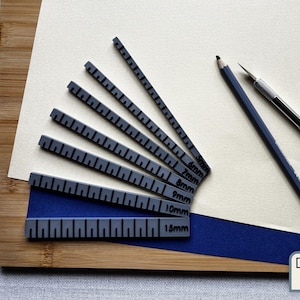 May include: A collection of gray rulers with black markings, ranging from 5mm to 15mm, arranged on a wooden surface. A blue pencil and a silver craft knife are also present. The rulers are on a white and blue background.