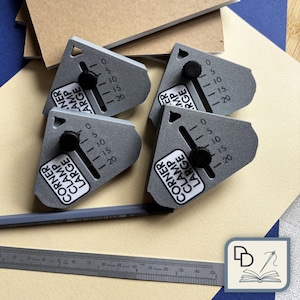May include: Four gray corner clamps with black knobs, labeled "CORNER CLAMP LARGE", are arranged on a light tan surface. Each clamp has a scale marked from 0 to 20. A pencil and a ruler are also visible.
