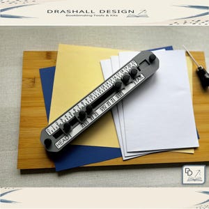 Op de afbeelding: Een grijze boekbindtool met een liniaal en verstelbare knoppen, gemarkeerd met "HEAD" en "TAIL". Het ligt op een houten oppervlak met wit, goud en blauw papier. De tekst "DRASHALL DESIGN Bookbinding Tools & Kits" staat bovenaan.