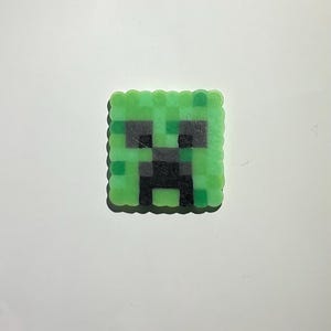 May include: A square-shaped pixel art piece crafted from fused plastic beads. The design features a green and black blocky face with two dark square eyes and a central dark mouth area, mimicking a classic video game creature character.
