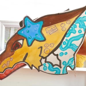 Può includere: Un ritaglio di testa di drago colorato, disegnato a mano. Il drago presenta sezioni marroni, gialle e bianche con accenti blu, tra cui una stella marina e linee ondulate. La bocca è rossa e il design include piccoli cerchi e rettangoli.