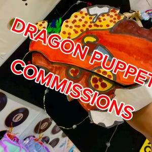 Op de afbeelding: Een handgemaakte drakenpop met een levendig oranje en geel kleurenschema met een gevlekt patroon. De pop heeft een opvallende kopvorm en is voorzien van de witte tekst DRAGON PUPPET COMMISSIONS in een vetgedrukt rood omlijnd lettertype.