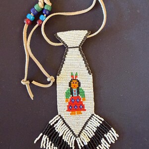 May include: Handmade beaded necklace featuring a Native American figure. The pendant is white with a black border and a beaded image of a person in a red dress and green top. The necklace has a leather cord and colorful beads.