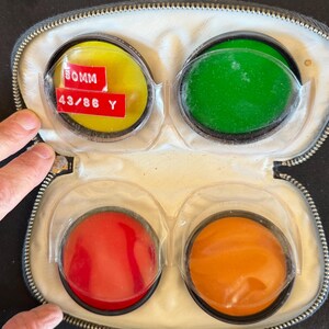 May include: A zippered carrying case containing four circular camera lens filters in yellow, green, red, and orange. The yellow filter features a red label with the white text 50MM and 43/86 Y. Each filter is held in a clear plastic sleeve.
