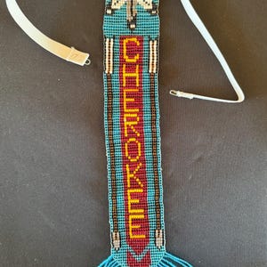 May include: A beaded headband featuring a turquoise background with a central red vertical panel displaying the word CHEROKEE in yellow letters. The design includes a moth motif at the top, three yellow stars, and a turquoise fringe with yellow beads.