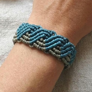 May include: A handmade macrame bracelet in shades of blue and gray is displayed on a wrist. The bracelet features a woven design. The image also shows a gold ring and dried flowers. The wooden sign says "Atelier Macramé - Handmade -."