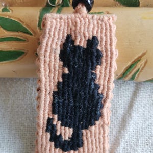 Op de afbeelding: Een perzik macramé sleutelhanger met een zwart katten silhouet. De sleutelhanger heeft een zilverkleurige sluiting en een zwarte kraal. De onderste rand heeft een franje detail. De achtergrond is een lichtgekleurde stof en een houten decoratief stuk.
