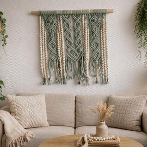 Peut inclure: Intérieur de salon avec un canapé beige, des coussins et une table basse en bois. Une tenture murale en macramé vert sauge et crème est suspendue au-dessus du canapé. Des plantes en pot et une lampe complètent l'ambiance.
