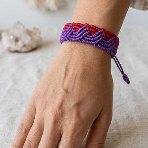 May include: A handmade macrame bracelet in shades of purple and red, worn on a wrist. The bracelet features a chevron pattern and adjustable closure. The background includes a neutral-toned textile and a decorative element.
