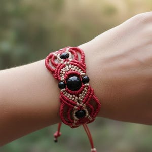 Könnte beinhalten: Rotes und beiges Makramee-Armband mit schwarzen Perlen. Das Armband hat ein gewebtes Design mit einem zentralen Muster aus schwarzen Perlen und einer roten Kordel. Das Armband ist mit einer roten Kordel gebunden.