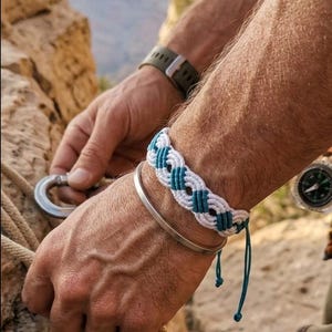 May include: A macrame bracelet with white and teal woven pattern. A silver bracelet and a green watch are also visible. The person is holding climbing gear, with a scenic background of a canyon.