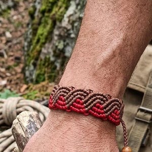 May include: A handmade macrame bracelet in shades of brown and red. The bracelet features a woven chevron pattern and a wooden bead closure. The image is set outdoors with a natural background, suggesting a rustic or bohemian style.