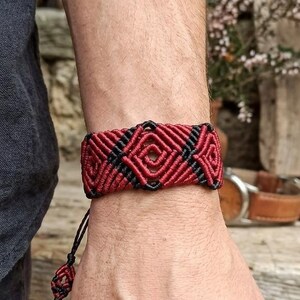 May include: A handmade macramé bracelet in red and black, with a geometric diamond pattern. The bracelet is approximately 2 cm wide and is worn on a wrist. The style is bohemian and suitable for everyday wear.