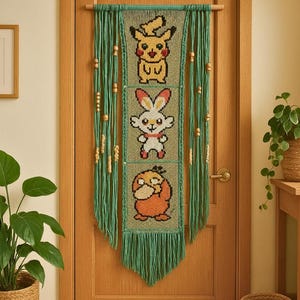 May include: A green macrame wall hanging featuring three panels with embroidered cartoon characters. The top panel shows a yellow character, the middle panel shows a white rabbit, and the bottom panel shows an orange character. Wooden beads and fringe complete the design.