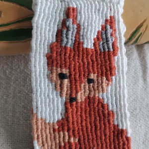 以下が含まれることがあります： 手作りの白とオレンジ色のキツネのキーホルダー。キツネはピクセル化されたデザインの織り糸で作られています。キーホルダーには、シルバーのリングと、緑色のビーズが付いた白い糸のループが付いています。