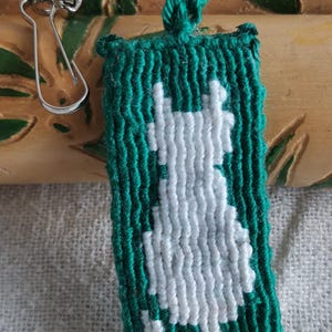 Op de afbeelding: Een handgemaakte groene en witte sleutelhanger met een zeepaardontwerp. Het zeepaardje is wit en de achtergrond is groen. De sleutelhanger heeft een zilveren sluiting en witte kralen. Het item is gemaakt van geweven garen.