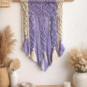 May include: A lavender and cream macrame wall hanging. This handmade textile art piece features detailed knotting patterns, tassels, and a wooden dowel. This bohemian-style decor item adds texture and colour to a room. The wall hanging is a decorative item.