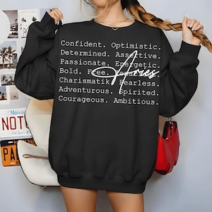 May include: Black sweatshirt with white text listing positive traits and the word "Aries" in a cursive font. The list includes words like "Confident," "Passionate," and "Adventurous." A red purse hangs in the background.