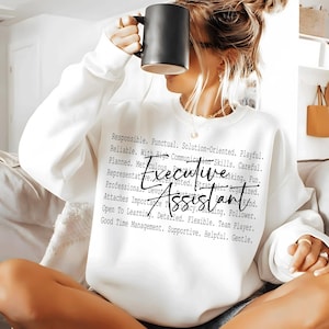 May include: White sweatshirt with the words "Executive Assistant" in a script font. The sweatshirt has a list of descriptive words in a smaller font. A black mug is held up to the face.