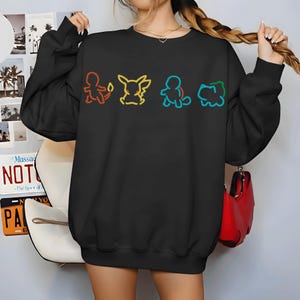 May include: Black sweatshirt featuring colorful line art of four cartoon characters. The characters are outlined in red, yellow, blue, and green. The sweatshirt is a crew neck style.