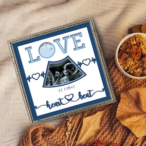 May include: A square silver-toned frame containing a white print with a blue border. The text reads LOVE AT FIRST HEARTBEAT, featuring a central ultrasound image of a fetus, a light blue clock icon, and dark blue heart-shaped EKG line graphics.