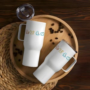 May include: Two white travel mugs with handles and lids, each adorned with a colorful line drawing of cartoon characters. The mugs are on a wooden tray with coffee beans and a woven placemat.