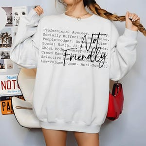 May include: A white long-sleeve sweatshirt featuring black text that reads: Professional Avoider, Low Key, Socially Buffering, Reclusive, People-Dodger, Awkwardologist, Social Ninja, Silent Observer, Ghost Mode, Energy-Preserving, Crowd Escape Artist, Selective Mute, Homebody Hero, Low-Volume Human, Anti-Social, and Not Friendly.