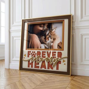 Puede incluir: Un cuadro cuadrado de pared con una fotografía de una persona sosteniendo un gato atigrado marrón y blanco. Debajo de la foto, el texto FOREVER IN MY HEART aparece en letras rojas negritas, decorado con una huella y pequeños corazones dorados.