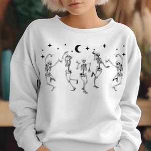 May include: A white long-sleeve sweatshirt featuring a black graphic print of five skeletons in various dancing poses. The design includes a crescent moon and several four-pointed stars positioned above the skeletons across the chest area.