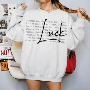 Könnte beinhalten: Ein weißes Langarm-Sweatshirt mit einem schwarzen Grafikdruck des Wortes Luck in einer großen Schreibschrift, überlagert mit einer Liste von Phrasen wie Shamrock Sparkle Magic, Lucky Charm, Kiss of Luck, Golden Streak und Leprechaun Gold.