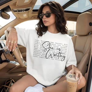 Op de afbeelding: Een wit T-shirt met korte mouwen voorzien van een blok zwarte tekst met diverse inspirerende woorden en de grotere cursieve tekst "She is Worthy" in het midden. Het shirt heeft een ronde halslijn en een ruimvallend, ontspannen model.