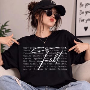 May include: A black short-sleeved t-shirt featuring white text listing autumn-themed words like Cozy Sweaters, Cinnamon, Scarves, Jeans, Flannel, Apple Cider, Pie, Pumpkin Patches, Haystack, Plaid, Giving Thanks, Harvest, Football, and the word Fall.